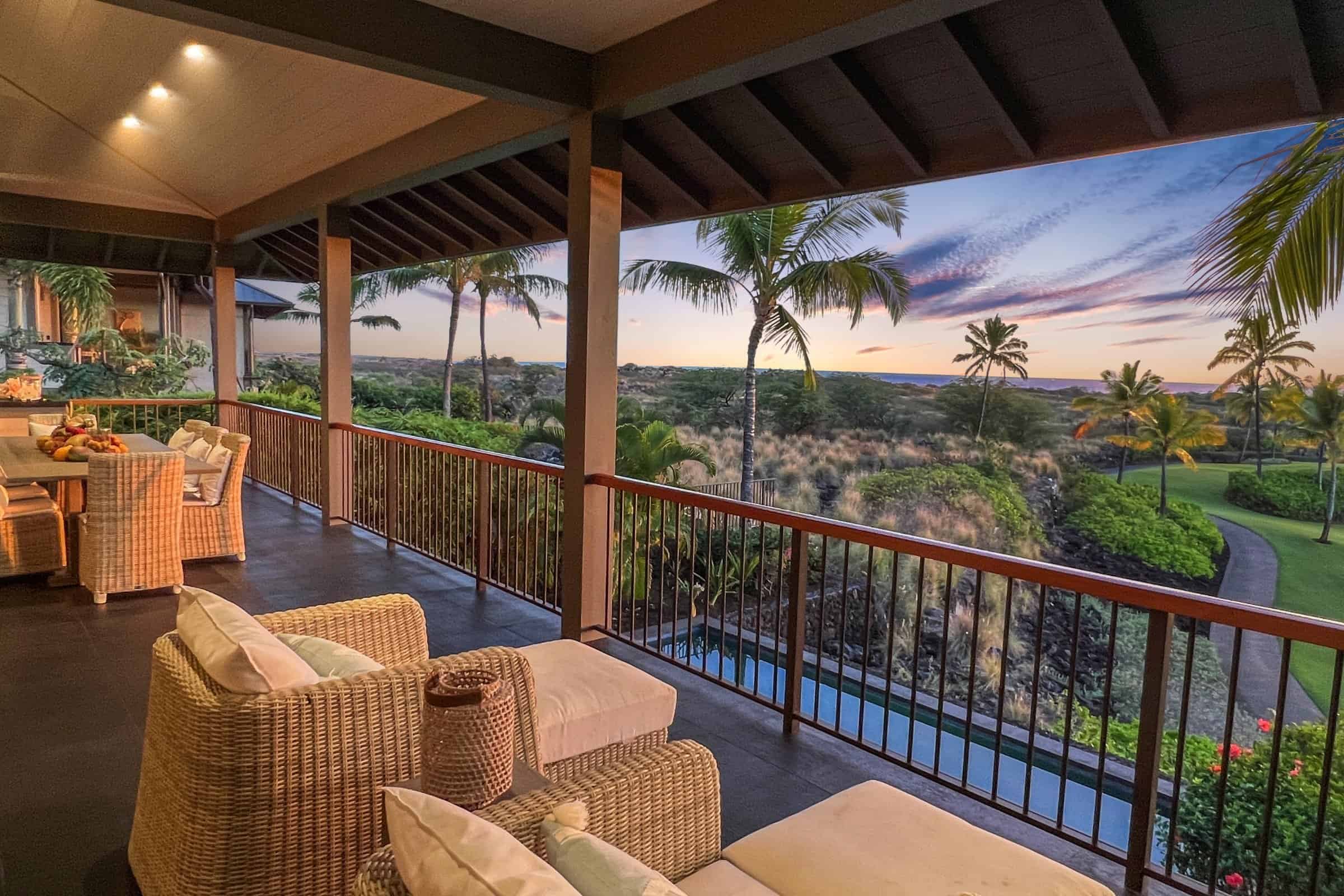 Covered lanai with wicker seating looking out over palms and lava at sunset in Kohanaiki.