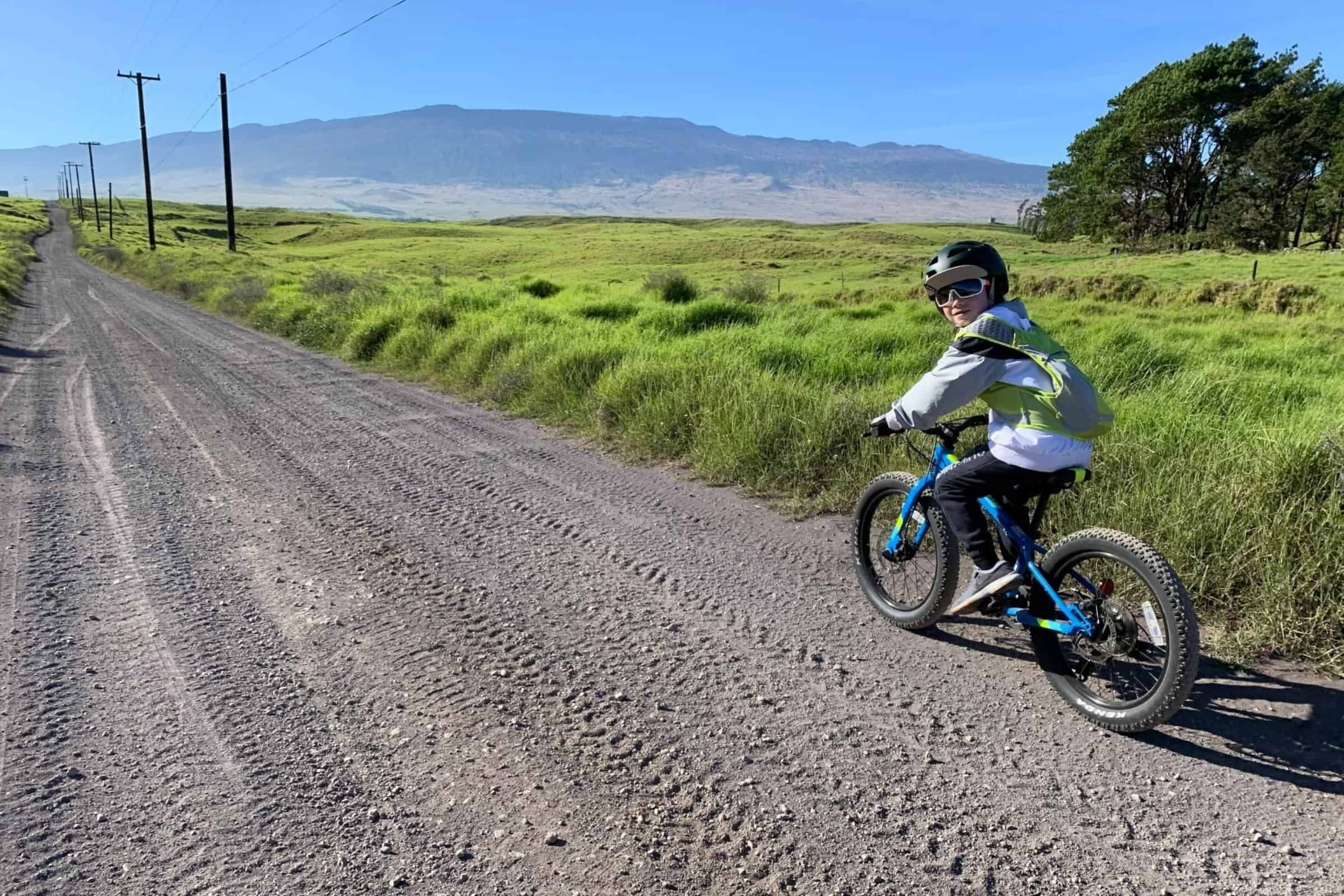Child riding a bike on a dirt road in rural Big Island Hawaii with lush greenery and distant mountai.