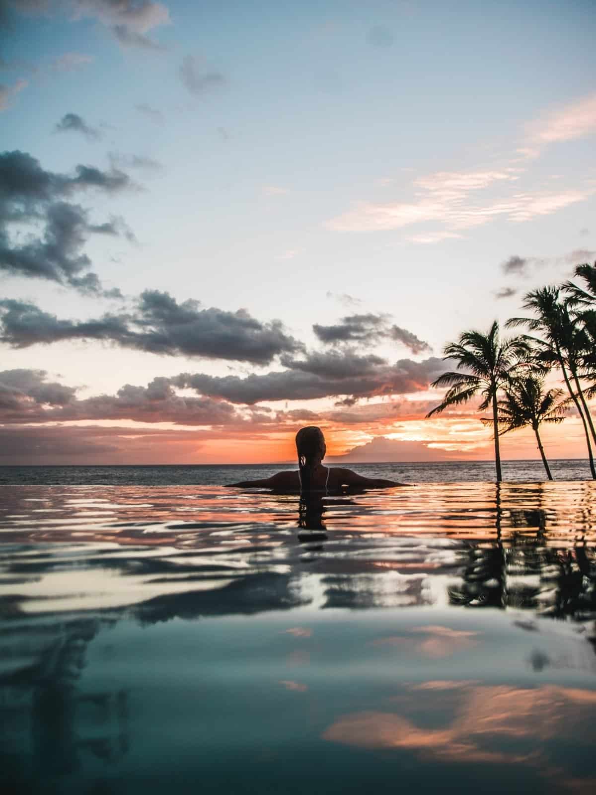 Woman in an infinity pool at sunset, palms silhouetted against the ocean.